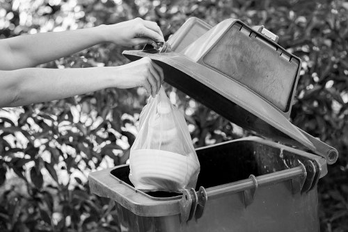 A person placing a plastic bag filled with household waste into a large, dark grey wheelie bin situated outdoors. The bin is positioned on a paved surface with a background of dense, leafy bushes, indicating a residential or commercial area in Harlesden, London. The lid of the bin is open, revealing a textured bin liner and other waste items inside. The person's hands are visible, holding the bag and the lid, with the scene illuminated by natural daylight. The overall setting suggests routine rubbish collection in an urban outdoor environment, pertinent to commercial waste removal services in the Harlesden area. The scene emphasizes the practical process of waste disposal, including the use of standard bin equipment often employed in local rubbish collection operations.