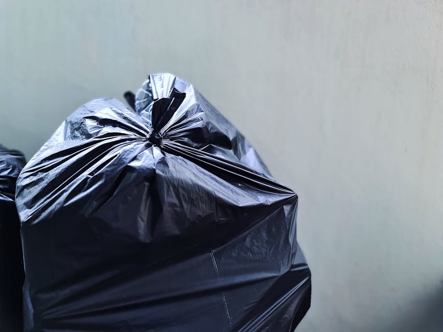 A pile of black plastic rubbish bags is gathered at the base of a green wooden building with weathered, painted shingle siding. The bags are stacked on a concrete sidewalk, near the bottom of a small 